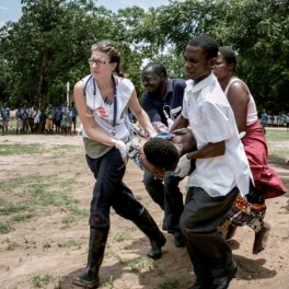 Eine Krankenschwester von Ärzte ohne Grenzen evakuiert mit weiteren unterstützenden Personen eine schwangere Frau über ein Feld in Richtung Hubschrauber.