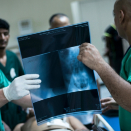 Zwei Chirurgen halten ein Röntgenbild in der Hand, hinter ihnen ein Patient auf einem Bett in der Notaufnahme.